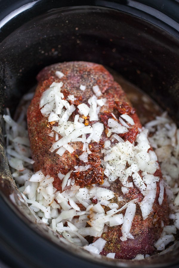Uncooked beef chuck roast, onions, and spices in slow cooker.