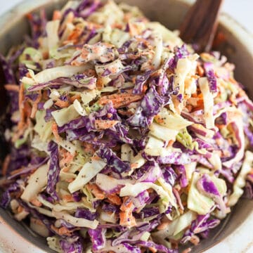 Red and green cabbage coleslaw in ceramic bowl with wooden spoon.