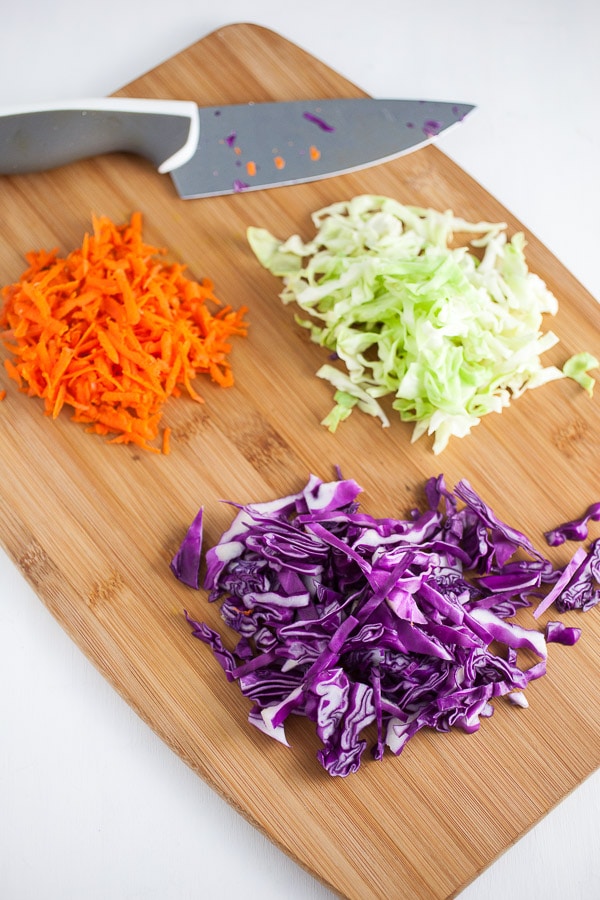 Shredded red and green cabbage and grated carrots on wooden cutting board with knife.