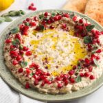 Mutabal eggplant dip garnished with pomegranate arils and fresh herbs on plate next to flatbread.