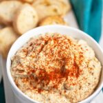 Baba ghanoush eggplant dip in white bowl garnished with smoked paprika next to toasted crostini on white tray.