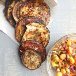 Mediterranean eggplant chips falling out of white package next to bowl of salsa.