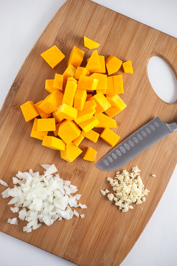Minced garlic and onions and peeled butternut squash cubes on wooden cutting board with knife.