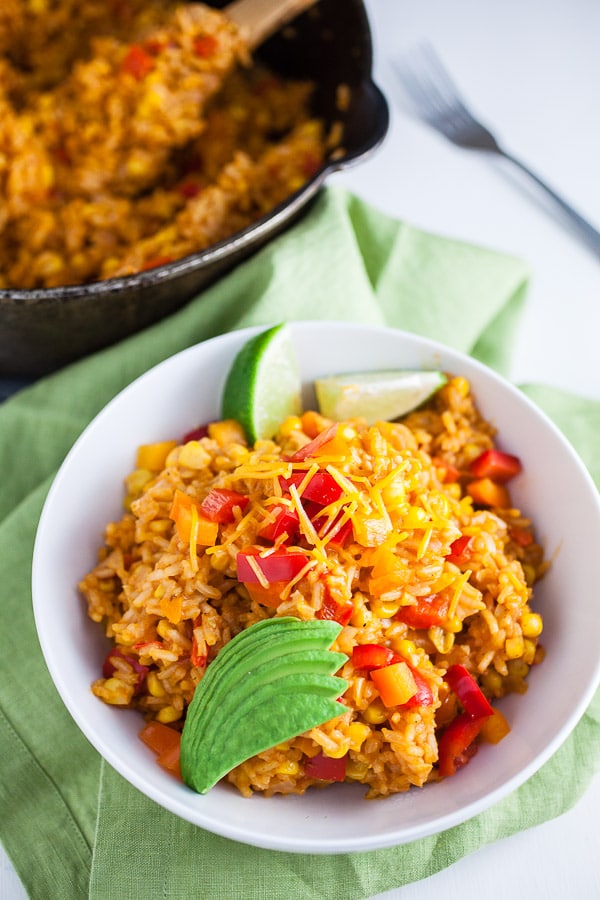 Cheesy Spanish rice with sliced avocado and lime wedges in white bowl next to cast iron Dutch oven.