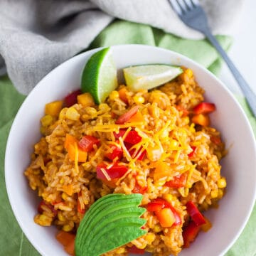 Cheesy Spanish rice with sliced avocado and lime wedges in white bowl.