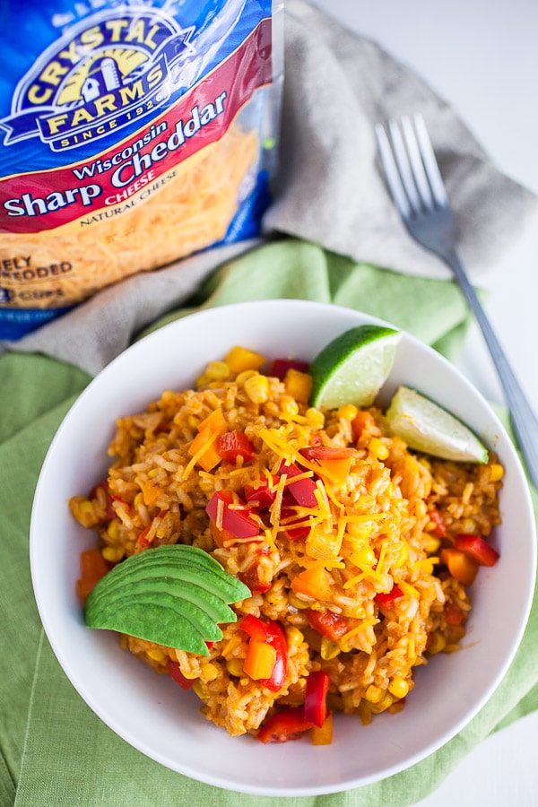 Spanish rice with sliced avocado and lime wedges in white bowl next to package of shredded cheese.