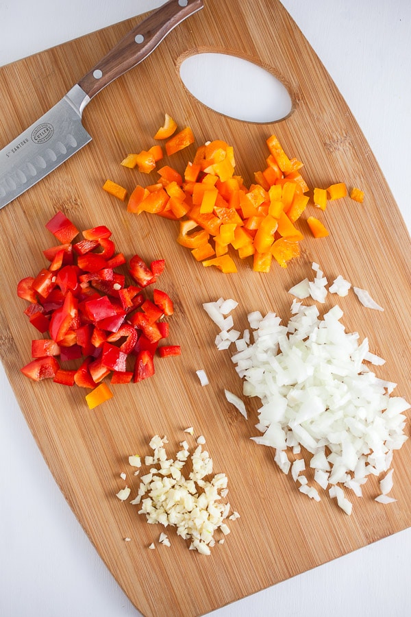 Minced garlic, onions, red and orange bell peppers on wooden cutting board with knife.