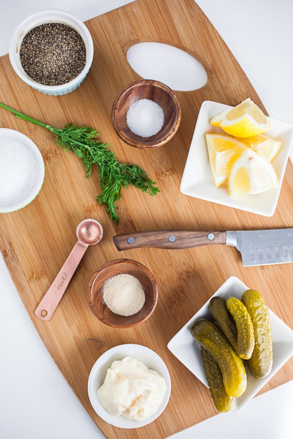 Mayo, pickles, salt, pepper, sugar, garlic powder, lemon wedges, and fresh dill on wooden cutting board.