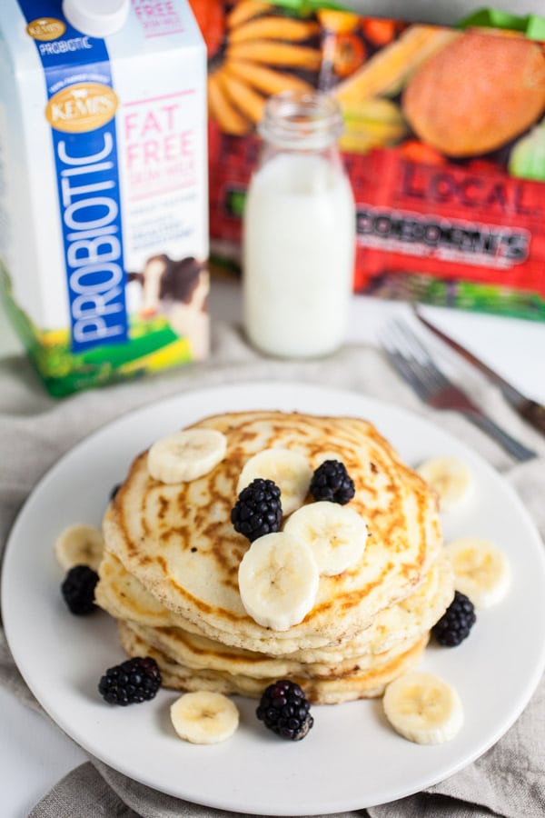 Stack of pancakes on white plate topped with bananas and blackberries next to reusable grocery bag and carton of milk.