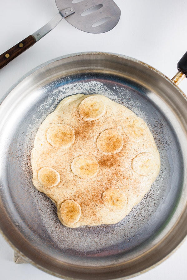 Pancake cooking in skillet with bananas and cinnamon.