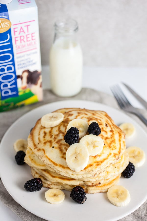 Stack of pancakes on white plate topped with bananas and blackberries next to carton of milk.
