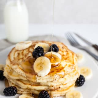 Maple syrup poured onto stack of old fashioned banana pancakes on white plate.