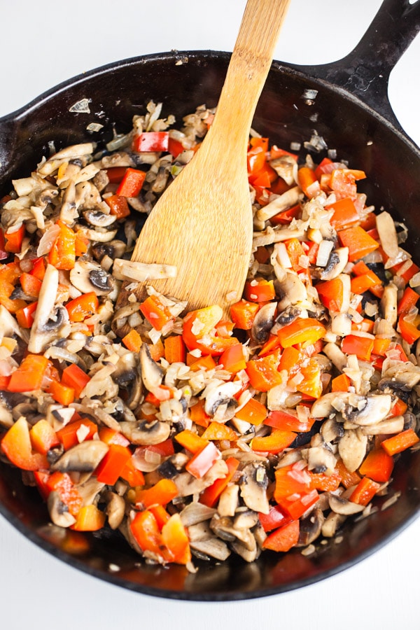 Garlic, onions, red bell peppers, and mushrooms sautéed in cast iron skillet with wooden spatula.