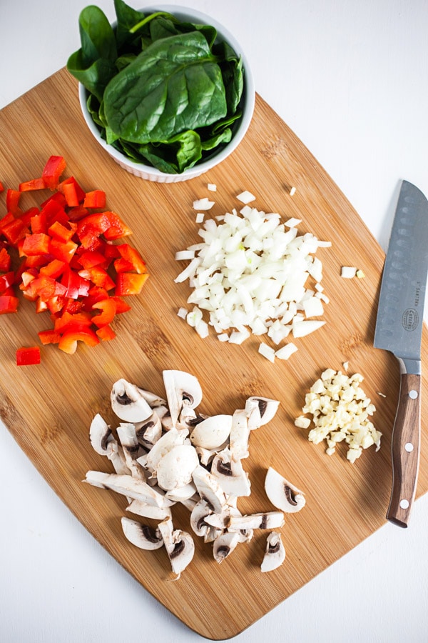 Minced garlic, onions, red bell peppers, mushrooms, and spinach on wooden cutting board with knife.