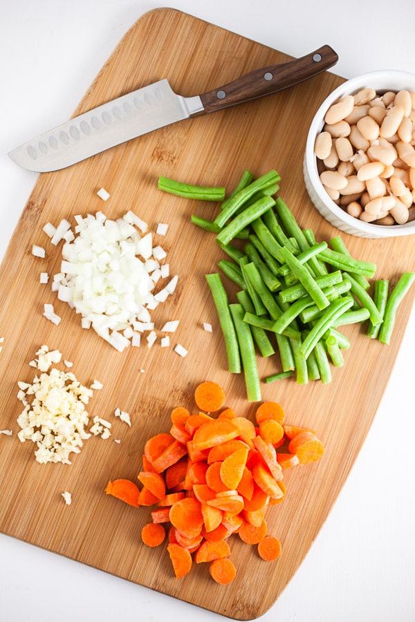 Minced garlic, onions, carrots, green beans, and white beans on wooden cutting board with knife.