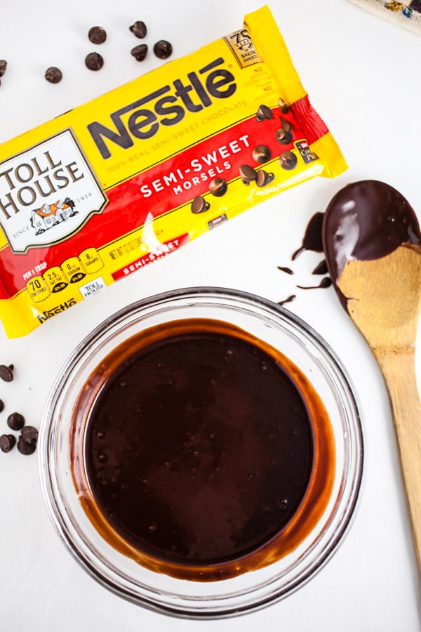 Chocolate ganache in small glass bowl next to wooden spoon and bag of chocolate chips.