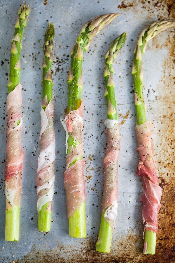 Uncooked prosciutto wrapped asparagus on metal baking sheet.