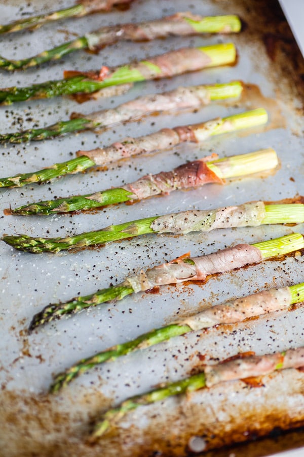 This Asparagus with Prosciutto and Creamy Lemon Tarragon Sauce is a tasty and decadent way to enjoy asparagus season! This prosciutto wrapped asparagus is drizzled with olive oil, baked in the oven, and roasted to perfection. It's served with a tangy and creamy lemon tarragon sauce. It makes a great easy appetizer recipe for holidays or parties! #asparagus #prosciutto #tarragon #sauce #appetizer Roasted asparagus with prosciutto on metal baking sheet.