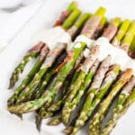 Prosciutto wrapped asparagus with creamy sauce on white serving plate with fork.
