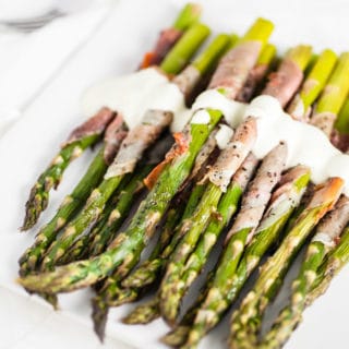 Prosciutto wrapped asparagus with creamy sauce on white serving plate with fork.