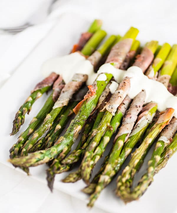 Prosciutto wrapped asparagus with creamy sauce on white serving plate with fork.