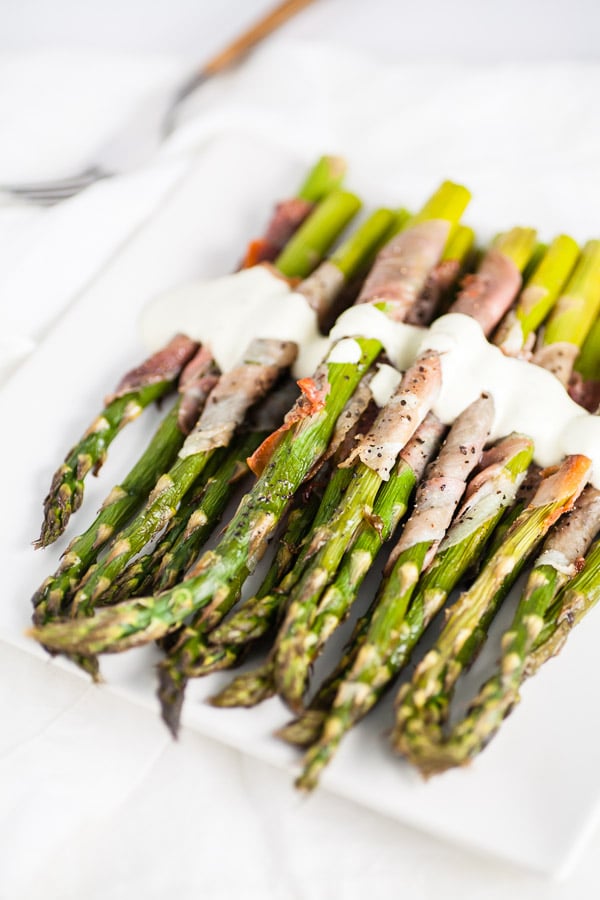 Prosciutto wrapped asparagus with creamy sauce on white serving plate with fork.