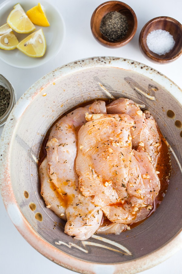 Chicken breasts marinating in ceramic bowl next to salt, pepper, and lemon wedges.