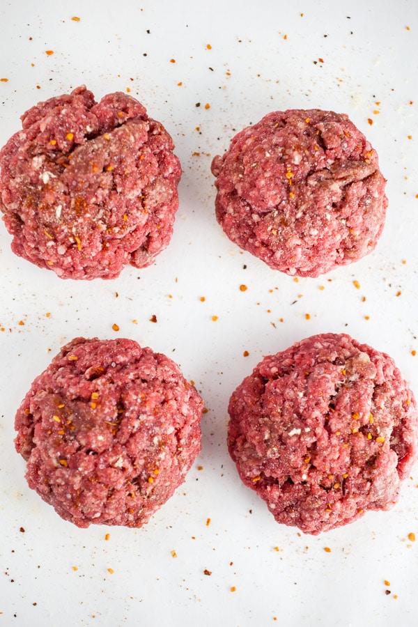 Raw ground beef patties with spices on white surface.