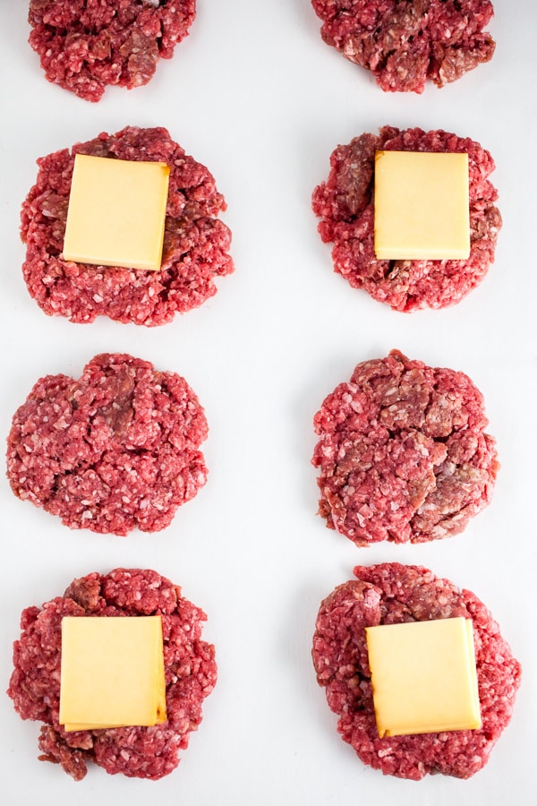 Uncooked beef patties topped with sliced cheese on white surface.