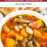 Vegetable soup in white bowl in front of pesto grilled cheese sandwich cut into halves.