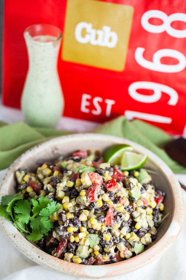 Black bean corn salad in ceramic bowl in front of glass container of dressing and reusable shopping bag.