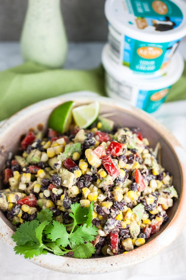 Black bean corn salad in ceramic bowl in front of sour cream containers and glass bottle of dressing.