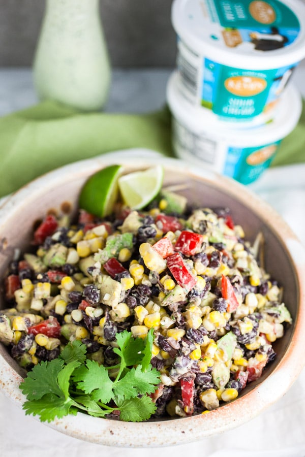 #ad This Southwest Black Bean and Corn Salad is easy to make, healthy, and fresh! It's great for summer parties and potlucks. It works as a salad, side dish, appetizer, or as a topping for burritos or tacos. It's made with a refreshing sour cream cilantro lime dressing. The Mexican flavors in this salad recipe are perfect for summer! @kempscows #southwest #salad #blackbeans #corn #cilantro #lime #sourcream Black bean corn salad in ceramic bowl in front of sour cream containers and glass bottle of dressing.