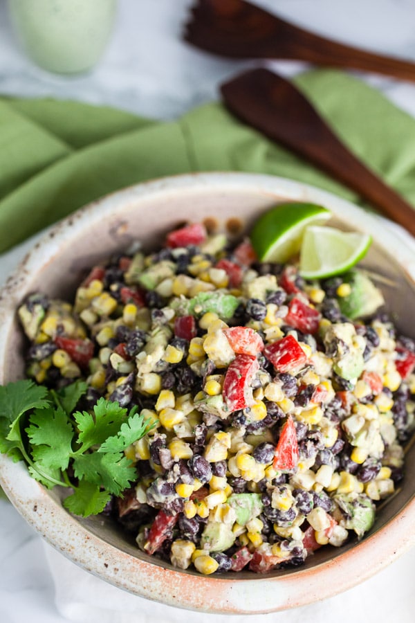 #ad This Southwest Black Bean and Corn Salad is easy to make, healthy, and fresh! It's great for summer parties and potlucks. It works as a salad, side dish, appetizer, or as a topping for burritos or tacos. It's made with a refreshing sour cream cilantro lime dressing. The Mexican flavors in this salad recipe are perfect for summer! @kempscows #southwest #salad #blackbeans #corn #cilantro #lime #sourcream Southwest black bean corn salad with fresh cilantro and lime wedges in ceramic bowl.