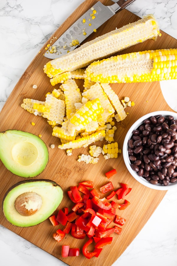Grilled corn, black beans, diced red bell peppers, and halved avocado on wooden cutting board with knife.
