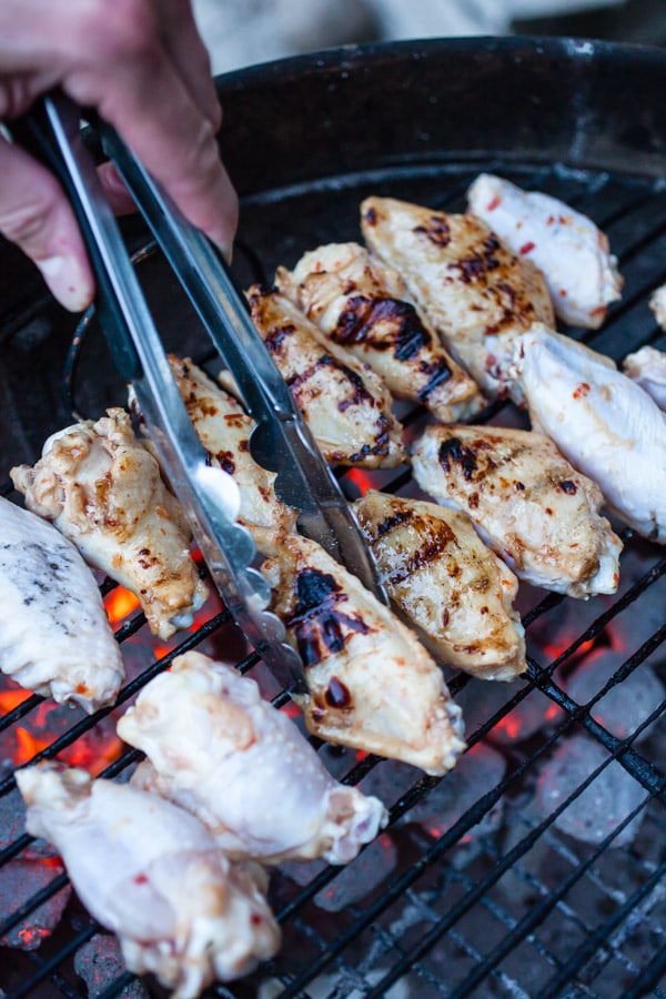 Hand holding metal tongs flipping chicken wings cooking on Weber grill.