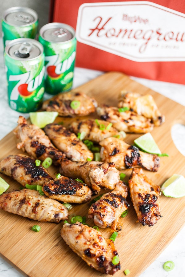 Grilled chicken wings on wooden cutting board in front of soda cans and reusable shopping bag.
