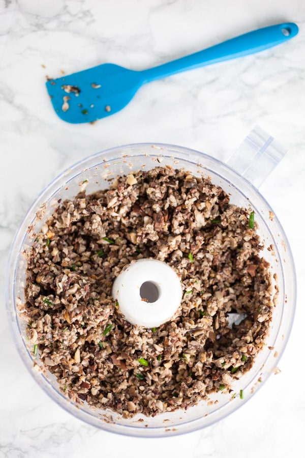 #AD MSG 4 21+ This Roasted Mushroom Tapenade Appetizer is the perfect simple and elegant recipe for your summer parties! Portobello and Crimini mushrooms are roasted on the grill and added to kalamata olives along with roasted garlic and walnuts. This healthy vegan and vegetarian dip is served on crostini. Serve with The Dreaming Tree wines for the perfect summer appetizer! #SeedOfAGreatSummer #mushrooms #tapenade #olives #roasted #appetizers Mushroom olive tapenade in food processor next to blue spatula.