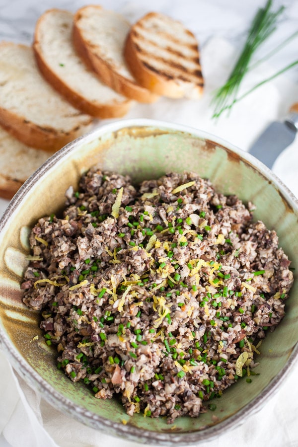 #AD MSG 4 21+ This Roasted Mushroom Tapenade Appetizer is the perfect simple and elegant recipe for your summer parties! Portobello and Crimini mushrooms are roasted on the grill and added to kalamata olives along with roasted garlic and walnuts. This healthy vegan and vegetarian dip is served on crostini. Serve with The Dreaming Tree wines for the perfect summer appetizer! #SeedOfAGreatSummer #mushrooms #tapenade #olives #roasted #appetizers Roasted tapenade in ceramic bowl with grilled crostini.