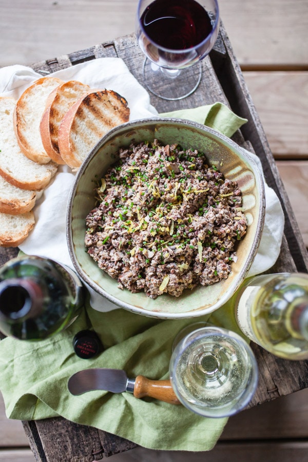 Mushroom tapenade in ceramic bowl with grilled crostini, wine glasses, and bottle of red and white wine on small table.