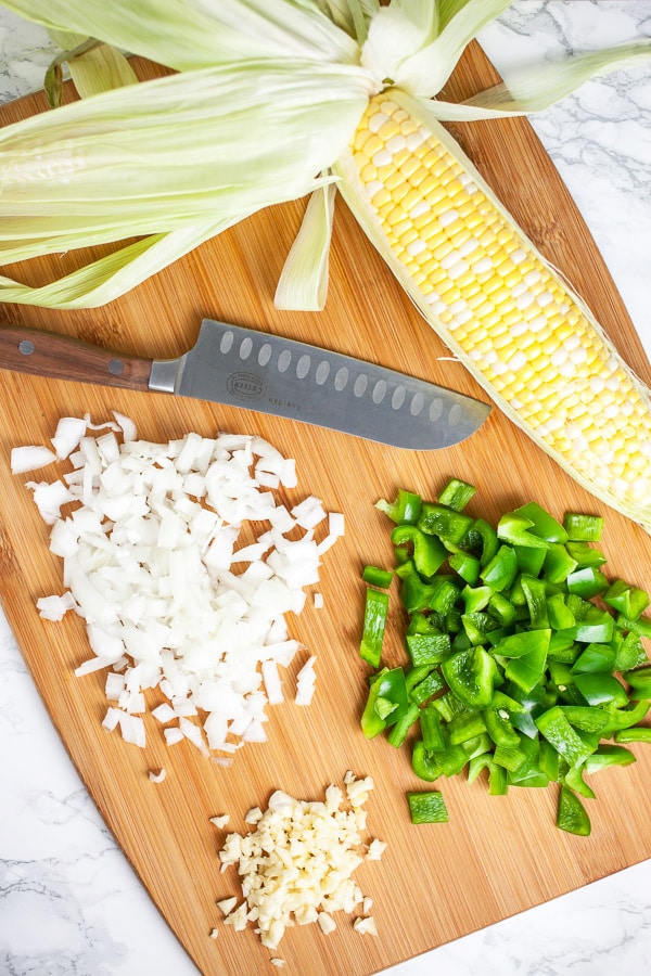 Minced garlic, onions, green bell peppers, and cob of fresh corn on wooden cutting board with knife.