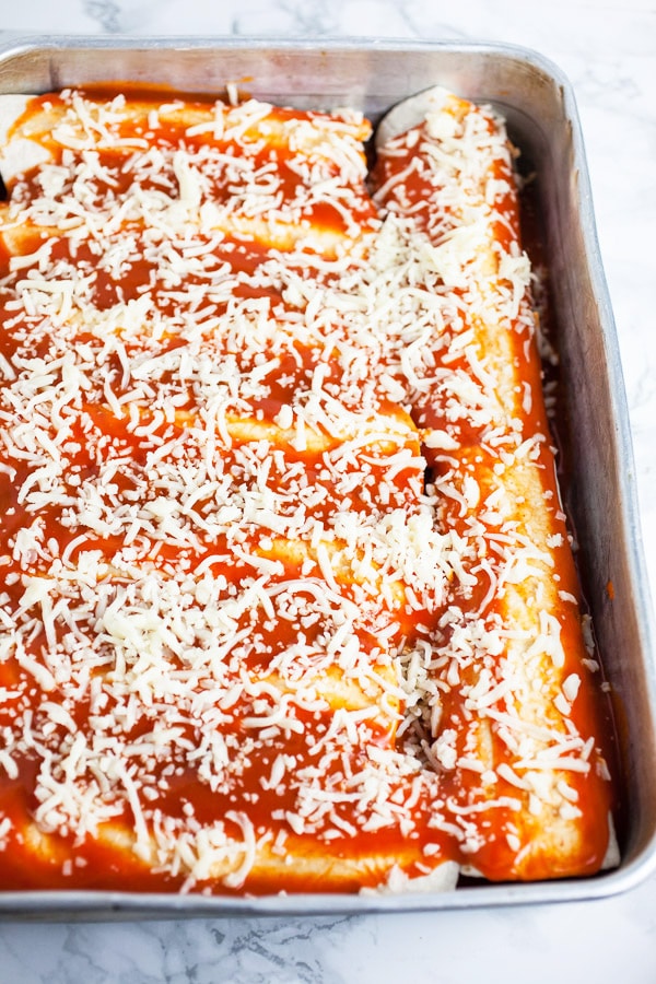 Unbaked enchiladas with enchilada sauce and shredded mozzarella cheese in cake pan.