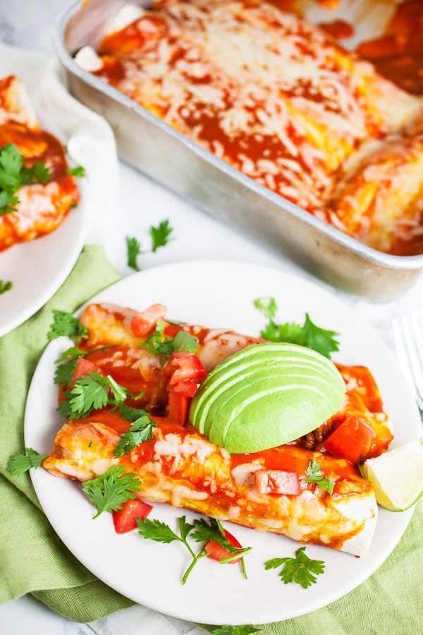 Cheesy ground beef enchiladas with fresh cilantro and avocado slices on white plates.