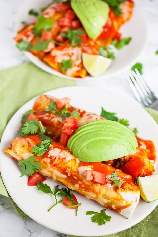 Cheesy ground beef enchiladas with fresh cilantro and sliced avocadoes on white plates.