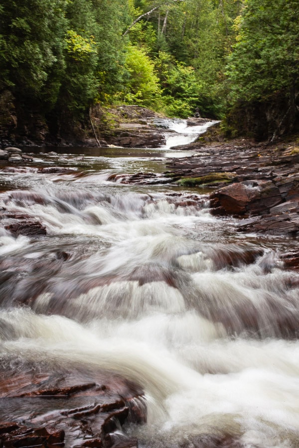 Read about our recent trip to Grand Maris in northern Minnesota! This charming small town sits on the shore of Lake Superior. It's home to an artist colony, numerous restaurants, and boasts miles of hiking trails. The natural beauty of the landscape is unbeatable. You'll often find picturesque sunsets and northern lights. Experience A Weekend In Grand Marais! #minnesota #greatlakes #lakesuperior #grandmarais #travel #vacation #explore Water running in river through the woods.
