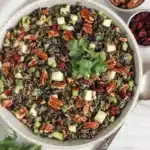 Wild rice salad garnished with parsley in serving bowl next to small bowls of cranberries and pecans.