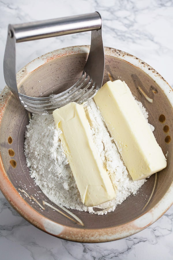 Flour and two sticks of butter in ceramic bowl with metal pastry cutter.