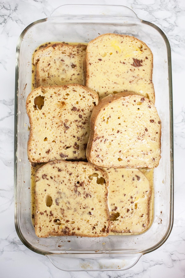 This Eggnog French Toast is a super easy breakfast recipe for Christmas morning and the holidays! Gluten free bread is soaked in a cinnamon egg mixture and then toasted until golden. It's topped with warm maple syrup and nutmeg. Everyone will love this French Toast! #breakfast #eggnog #christmas #glutenfree Pieces of bread soaking in egg mixture in glass baking dish.