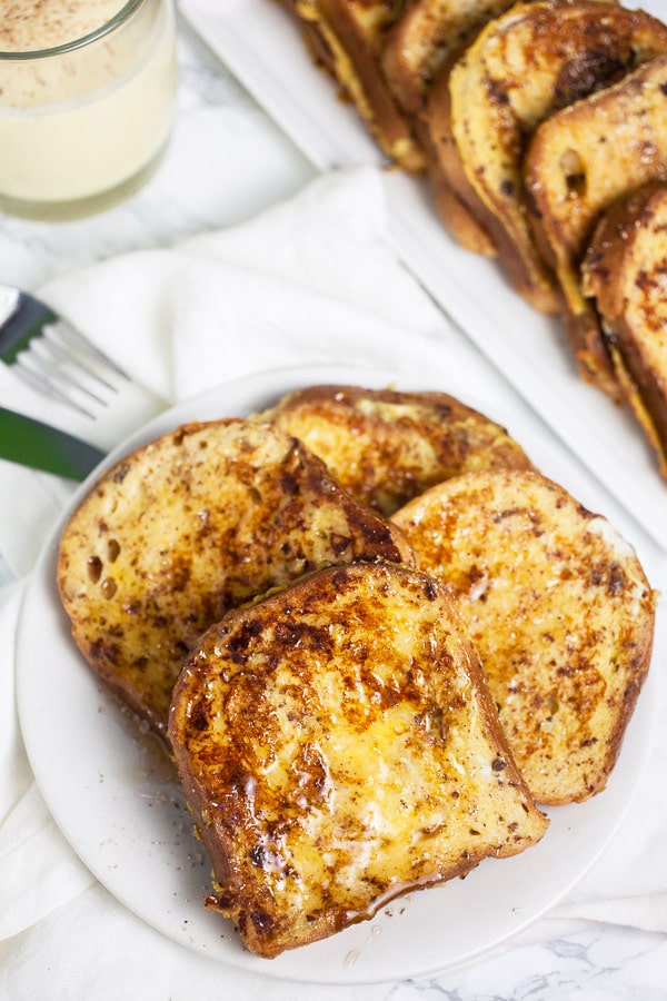 This Eggnog French Toast is a super easy breakfast recipe for Christmas morning and the holidays! Gluten free bread is soaked in a cinnamon egg mixture and then toasted until golden. It's topped with warm maple syrup and nutmeg. Everyone will love this French Toast! #breakfast #eggnog #christmas #glutenfree French toast with maple syrup on white plate with glass of eggnog.