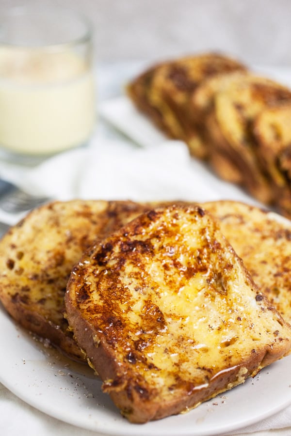 This Eggnog French Toast is a super easy breakfast recipe for Christmas morning and the holidays! Gluten free bread is soaked in a cinnamon egg mixture and then toasted until golden. It's topped with warm maple syrup and nutmeg. Everyone will love this French Toast! #breakfast #eggnog #christmas #glutenfree French toast with maple syrup on white plate with glass of eggnog.